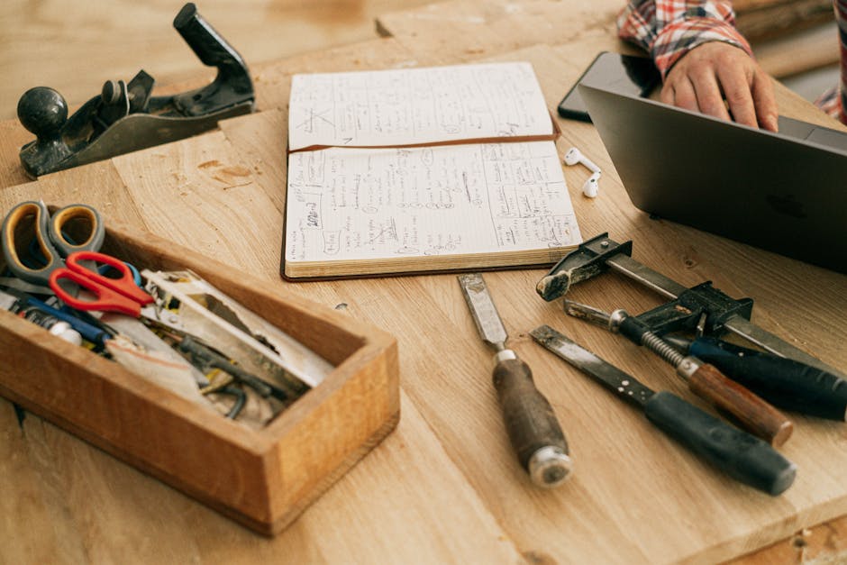 A workspace featuring woodworking tools, a notebook, and a hand on a laptop, symbolizing creativity and craftsmanship.
