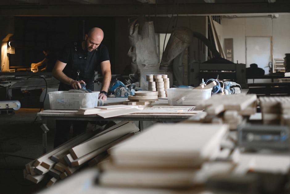 Concentrated male carpenter polishing wooden detail at table while working in professional joinery with abundance of wooden planks and special equipment