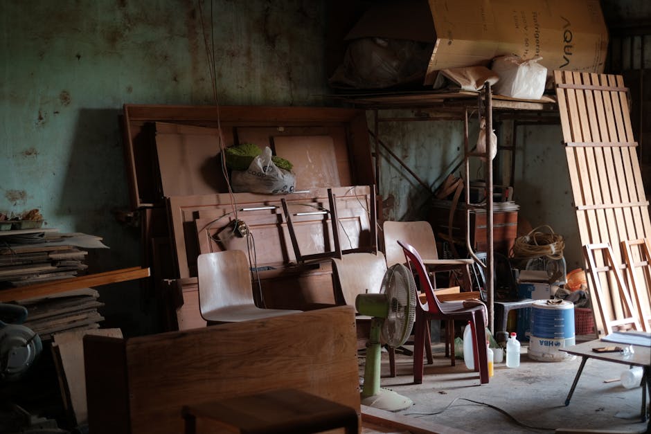 A cluttered workshop filled with wooden furniture, tools, and various items in natural lighting.