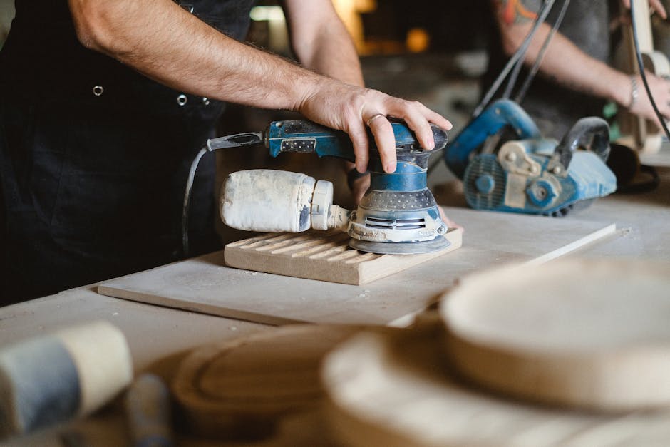Unrecognizable professional male carpenter using random orbital sander while polishing wooden board during work at workbench in joinery studio with tools