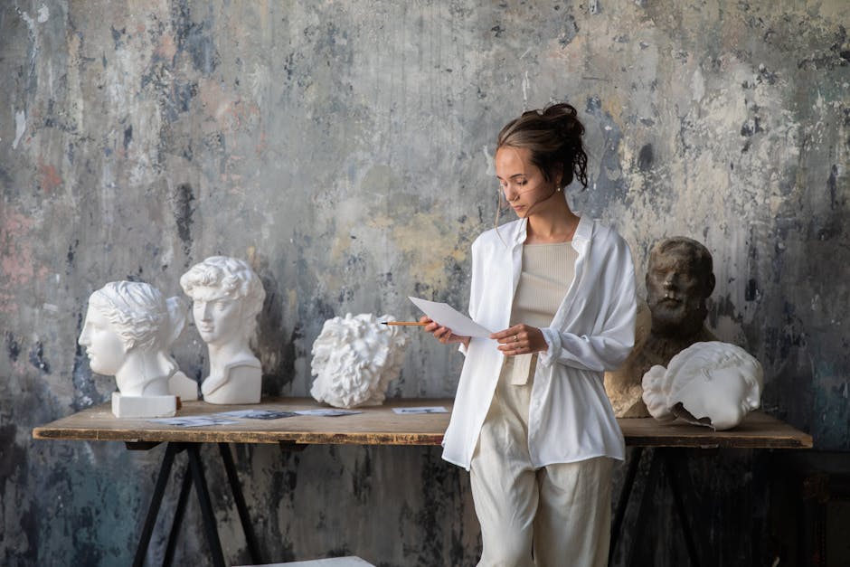 Female artist in workshop with classical marble busts, examining sketches.